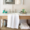 White Ines guest towel draped over a marble and wood bathroom vanity, with makeup accessories in green glass jars.