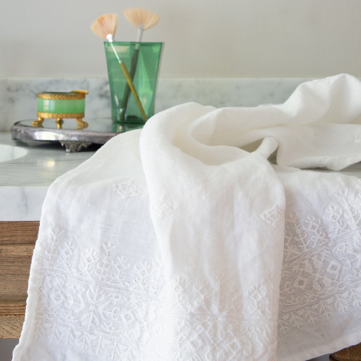 Ines Guest Towel | White | embroidered linen guest towel draped over a marble bathroom counter with green glass accessories in the background.