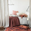 Ines Blanket | Ines throw blanket in mahogany, layered on a rumpled, blue, brown, and pink toned bed in linen and charmeuse, with a geometric rug and wrought-iron room accents - side view.