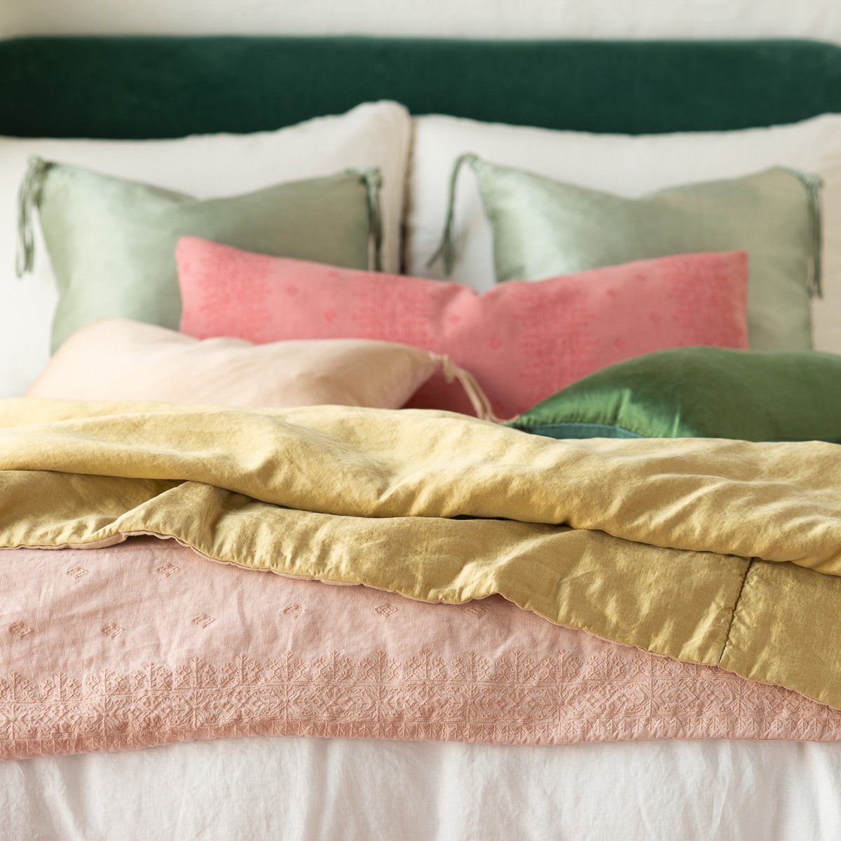 Ines Blanket |  Ines throw blanket in rouge, layered under green, pink, and gold tones on a white bed, - cropped end of bed view.