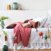 Linen bed skirt in white, layered on an mostly white bed with rich pink and brown accents, and surrounded by colorful vases - side view.