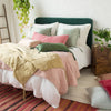 Shimmery Taline throw pillows and blanket in green and gold tones are paired with pink embroidered linen, with colorful rug, green plants, and warm wood accents - three-quarter angle.