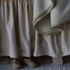 Harlow Crib Skirt | cotton velvet crib skirt in a neutral tone shown close up with the corner of a draped blanket visible along with tan-colored booties on a white-washed floor.