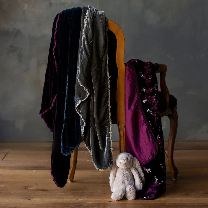 Lynette Baby Blanket | Silk velvet baby blankets in deep jewel tones draped on the back and arm of a chair with a small stuffed bunny on a medium wood floor. 