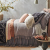 Linen Whisper bed skirt in moonlight, shown with matching duvet cover and shams, and layered with accessory pieces in rouge and parchment - side view.