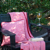 Adele Blanket | matching organic cotton damask throw blanket and 18" square pillow in Poppy on an outdoor chair with a glass of white wine on the armrest and two paperback books on the seat. Greenery is visible behind the chair.