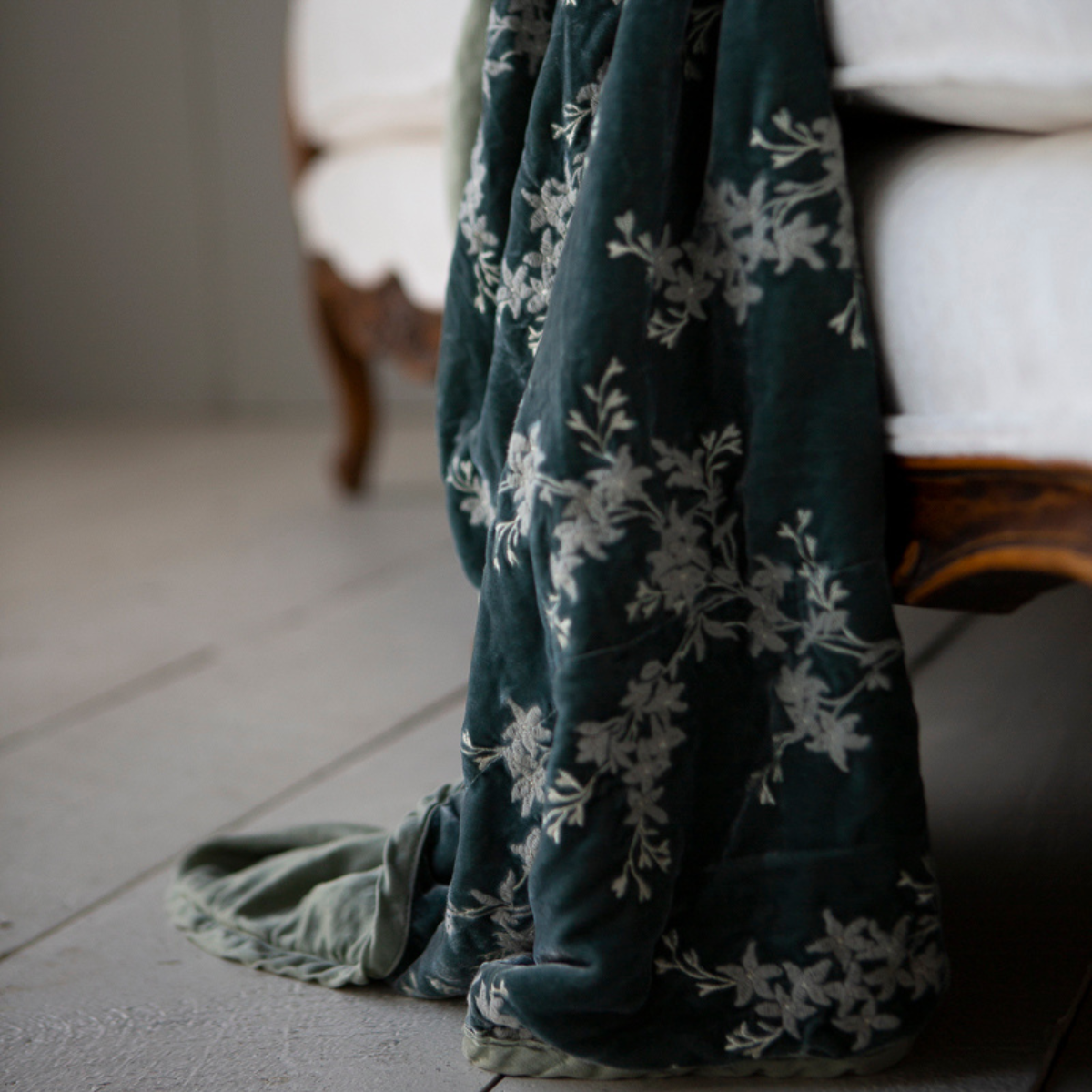 Lynette Blanket | Eucalyptus | close up of embroidered silk velvet with charmeuse trim and back, hanging off of the edge of a white couch sofa with wood detail.