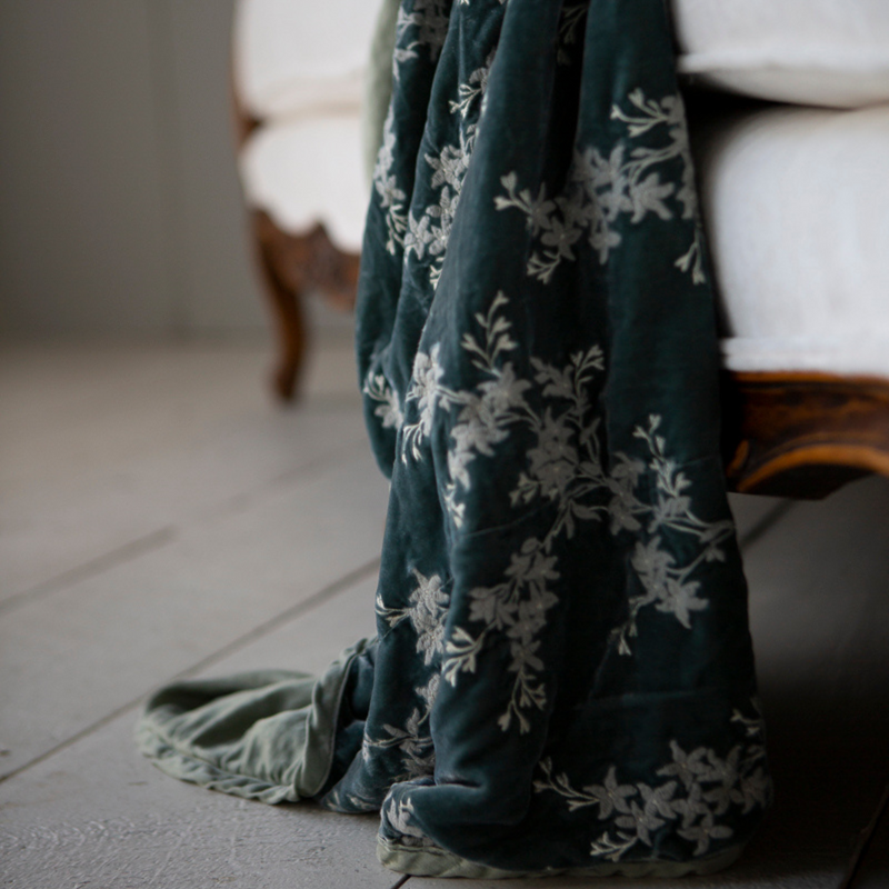 Lynette Blanket | Eucalyptus | close up of embroidered silk velvet with charmeuse trim and back, hanging off of the edge of a white couch sofa with wood detail.