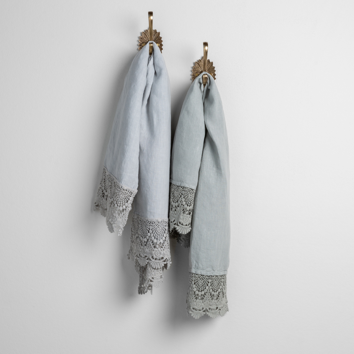 Mattine Guest Towel | cloud and eucalyptus linen guest towels with mattine lace hanging from decorative towel hooks against a plain white wall. 