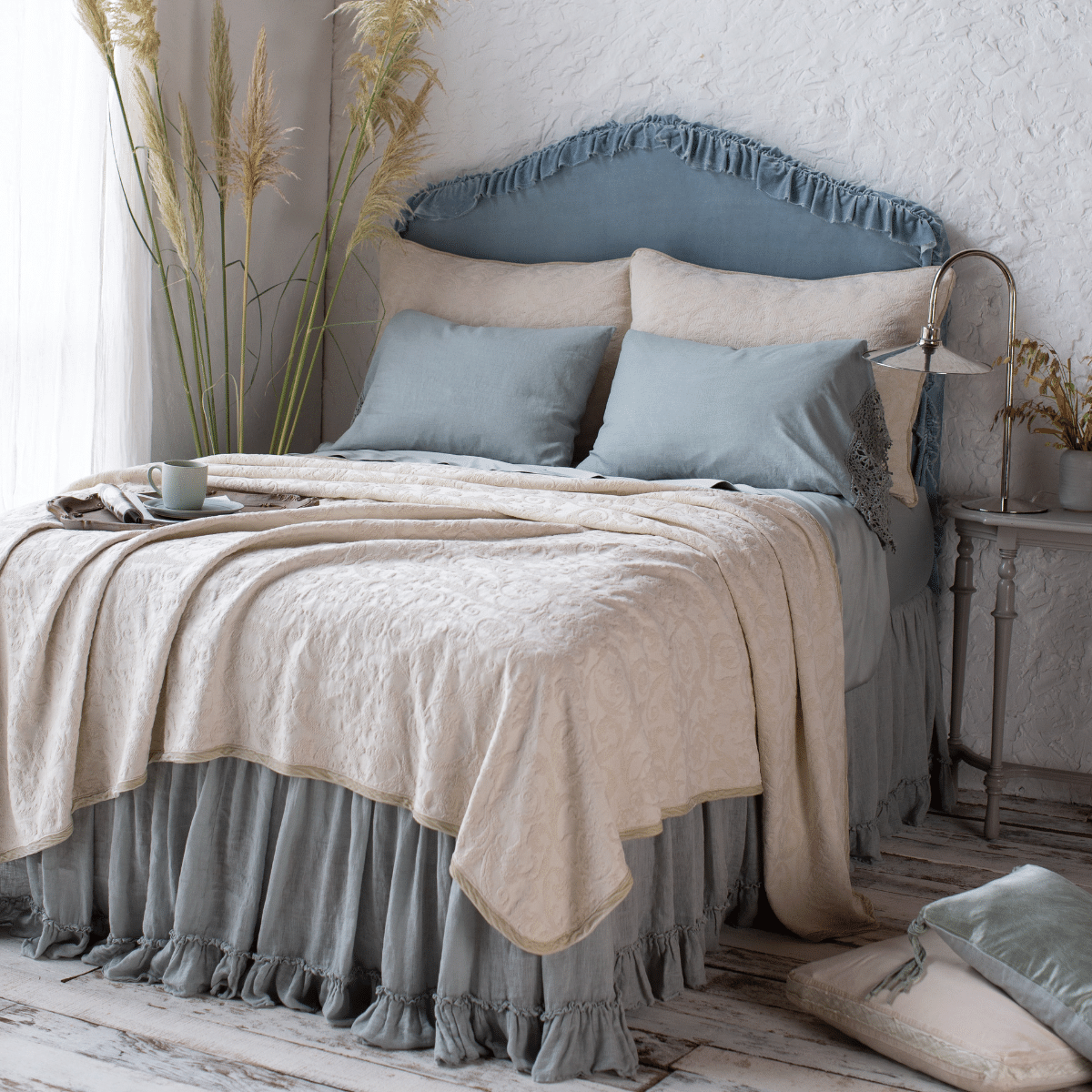 The Adele coverlet in parchment, shown with matching shams and Bria sheets in eucalyptus. Shot at an angle from the foot of the bed, the coverlet is beautifully rumpled over a ruffled bed skirt. The room in styled on distressed wood floors with dried greenery and natural light.