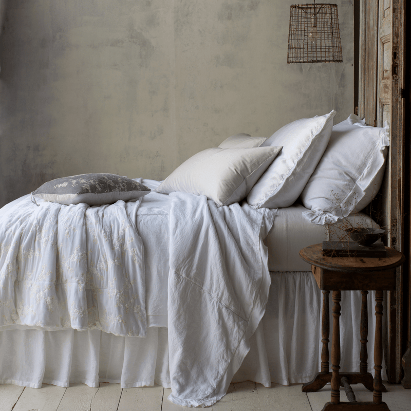 Lynette Blanket | White | Lynette throw blanket in white on a white and sterling bed in rumpled layers of linen, cotton, and silk charmeuse, against a  grey background and moody lighting - side view.