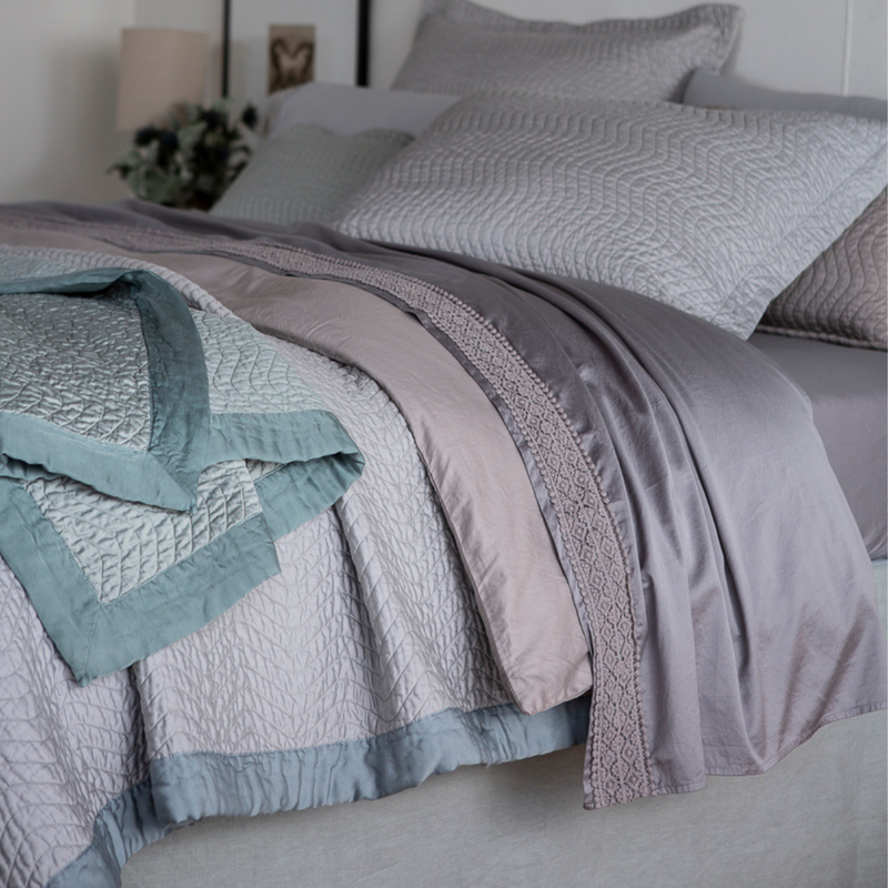 Cirillo Coverlet | a close up of the side edge of a bed dressed in cotton sateen. The shams, coverlet and throw blanket are quilted and the unquilted duvet cover and flat sheets are also visible. There is a band of cotton lace on the flat sheet's cuff.
