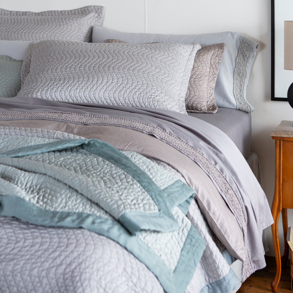 [allvariants]: Cirillo shams stacked at the head of the bed with other sleeping pillows and throw pillows. A flat sheet with lace  is visible, pulled back over the duvet cover and coverlet - the throw blanket at the end of the bed is intentionally layered haphazardly to show satin binding detail against the quilt pattern. 