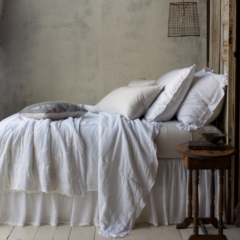 Bria Fitted Sheet | Sterling Bria sheets contrast against a mostly white bed, shown from the side. The lighting is moody and colors are muted, emphasizing the variety of cotton, linen, and silk textures.
