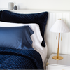 White Bria fitted sheet on a neatly made bed shown from close up three quarter angle. Layered with deep blue silk velvet quilted shams and coverlet, it showcases a minimalist and gender neutral look.