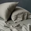 Rumpled Bria flat sheet in eucalyptus in the foreground, with matching sleeping pillows angled asymmetrically against a moody dark background.