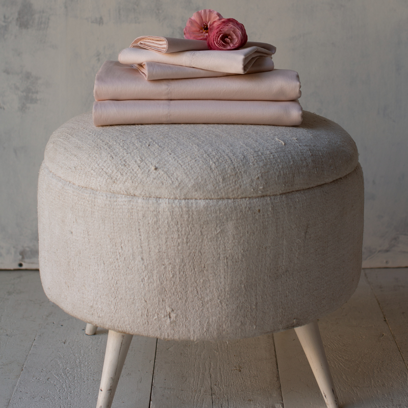 Bria Flat Sheet | Pearl | Soft pink Bria sheets and pillowcases, neatly folded and stacked on a light colored ottoman. Shown from the side, two dark pink flowers rest on top.