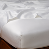 Bria Flat Sheet | White | Cotton sateen flat sheet draped over matching fitted sheet. Shown from the top corner, the flat sheet is rumpled, highlighting the shine of the fabric.