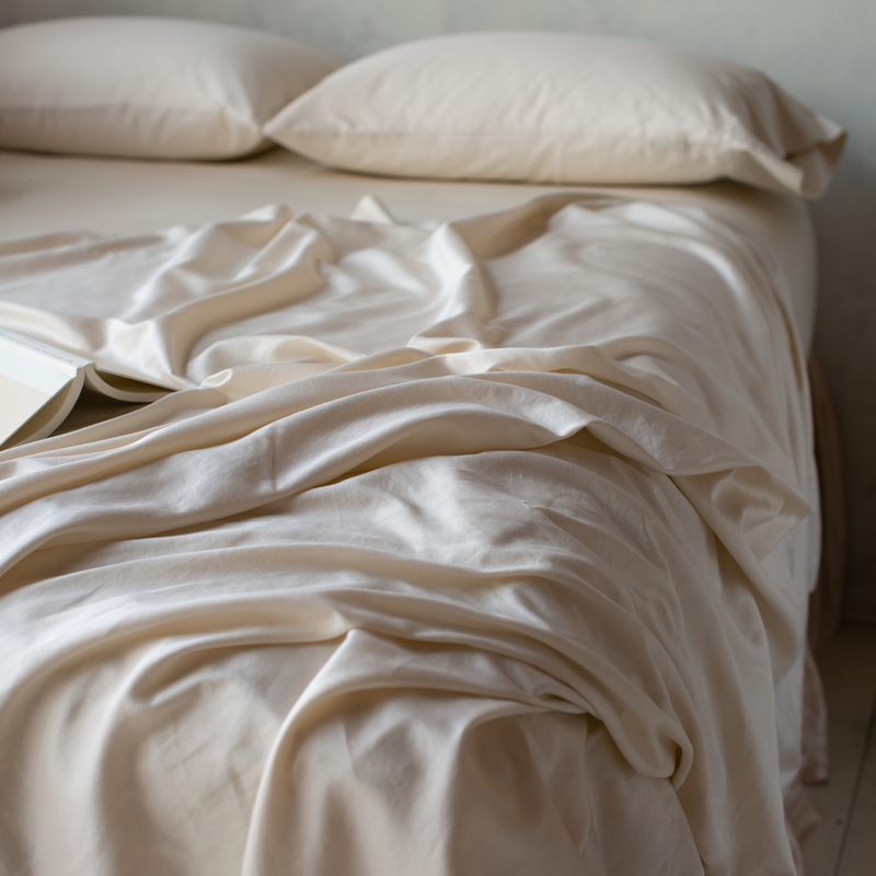 Bria Pillowcase (Single) | Parchment | Bria sleeping pillows in parchment, laid flat at the head of the bed, shown from three quarter angle. Matching sheets emphasize the sheen of the fabric.