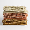 Carmen Blanket | silk velvet throw blankets folded in stack of three in warm tones against a white background, peitie ruffle trim detail shown bordering visible edges.