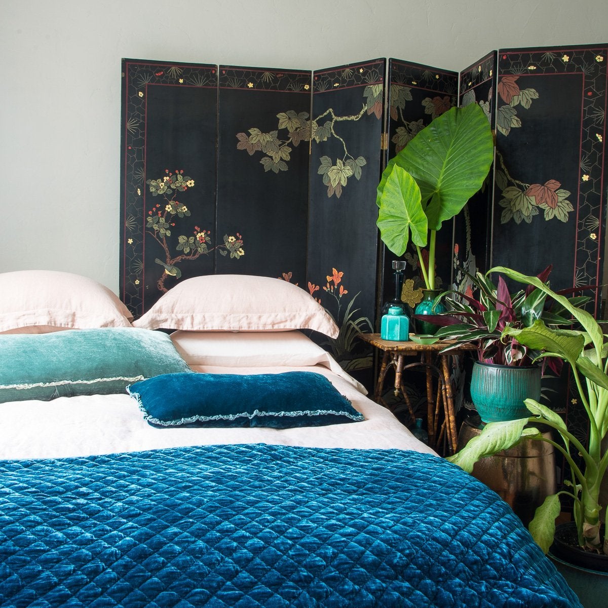 On a pearl pink linen bed, Carmen throw pillows in contrasting blue-green tones lay flat, highlighting the charmeuse, petite, raw-edge ruffle. Styled with on-tone ceramics and big green plants in front of a dark floral room divider.