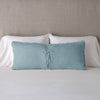 Carmen Throw Pillow | Cloud | Silk velvet lumbar pillow with petite ruffle leaning upright and backwards against neutral bedding and headboard, showcaseing the silk charmeuse tie closures.