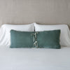 Carmen Throw Pillow | Eucalyptus | Silk velvet lumbar pillow with petite ruffle leaning upright and backwards against neutral bedding and headboard, showcaseing the silk charmeuse tie closures.