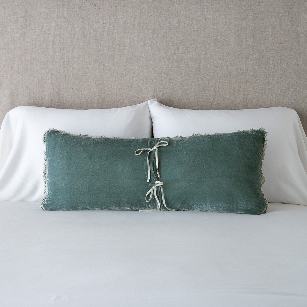 Carmen Throw Pillow | Eucalyptus | Silk velvet lumbar pillow with petite ruffle leaning upright and backwards against neutral bedding and headboard, showcaseing the silk charmeuse tie closures.