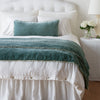 Carmen Throw Pillow | Eucalyptus | Silk velvet lumbar pillow with petite ruffle and throw blanket shown on an all white, neatly made bed - foot of bed angle.
