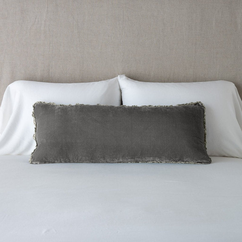 Carmen Throw Pillow | Fog | Silk velvet lumbar pillow with petite ruffle, leaning upright against neutral bedding and headboard.