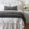 Carmen Throw Pillow | Fog | Silk velvet lumbar pillow with petite ruffle and throw blanket shown on an all white, neatly made bed - foot of bed angle.