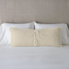 Carmen Throw Pillow | Parchment | Silk velvet lumbar pillow with petite ruffle leaning upright and backwards against neutral bedding and headboard, showcaseing the silk charmeuse tie closures.
