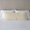 Carmen Throw Pillow | Parchment | Silk velvet lumbar pillow with petite ruffle leaning upright and backwards against neutral bedding and headboard, showcaseing the silk charmeuse tie closures.