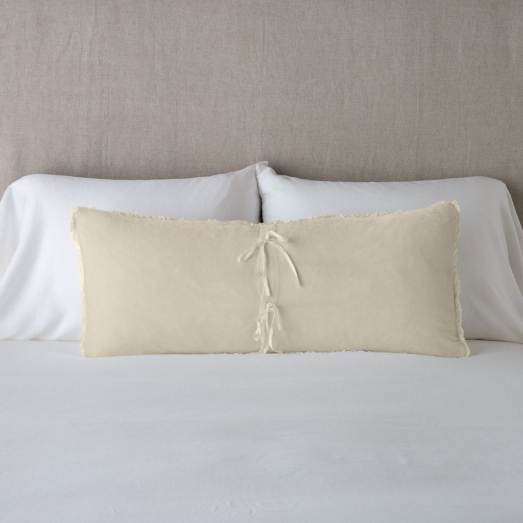 Carmen Throw Pillow | Parchment | Silk velvet lumbar pillow with petite ruffle leaning upright and backwards against neutral bedding and headboard, showcaseing the silk charmeuse tie closures.