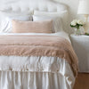 Carmen Throw Pillow | Pearl | Silk velvet lumbar pillow with petite ruffle and throw blanket shown on an all white, neatly made bed - foot of bed angle.