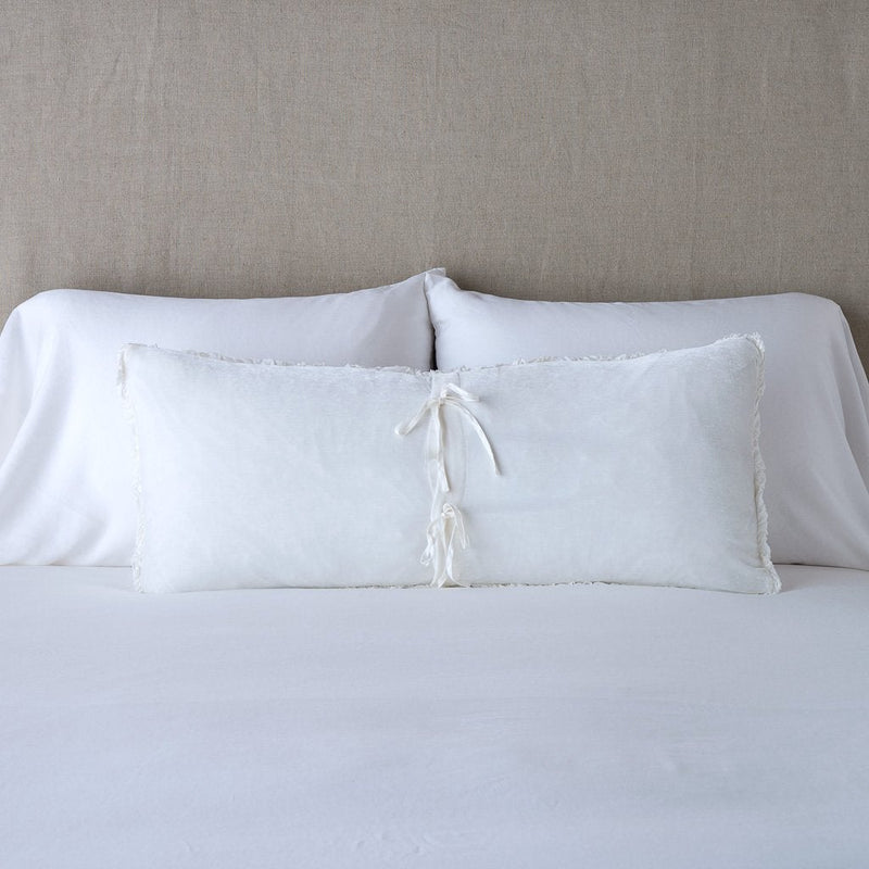 Carmen Throw Pillow | White | Silk velvet lumbar pillow with petite ruffle leaning upright and backwards against neutral bedding and headboard, showcaseing the silk charmeuse tie closures.