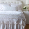 Carmen Throw Pillow | White | Silk velvet lumbar pillow with petite ruffle and throw blanket shown on an all white, neatly made bed - foot of bed angle.