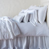 Carmen Throw Pillow | White | Silk velvet lumbar pillow with petite ruffle on an all white, neatly made bed with matching accessory pieces - side view.
