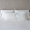Carmen Throw Pillow | Winter White | Silk velvet lumbar pillow with petite ruffle leaning upright and backwards against neutral bedding and headboard, showcaseing the silk charmeuse tie closures.