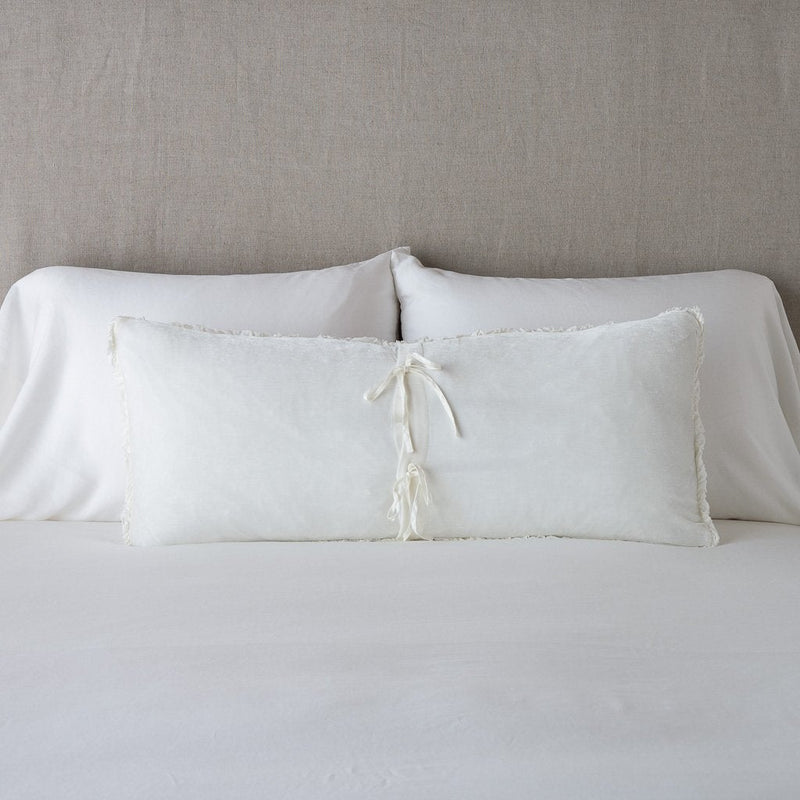 Carmen Throw Pillow | Winter White | Silk velvet lumbar pillow with petite ruffle leaning upright and backwards against neutral bedding and headboard, showcaseing the silk charmeuse tie closures.