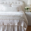 Carmen Throw Pillow | Winter White | Silk velvet lumbar pillow with petite ruffle and throw blanket shown on an all white, neatly made bed - foot of bed angle.
