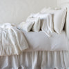 Carmen Throw Pillow | Winter White | Silk velvet lumbar pillow with petite ruffle on a monochromatic, neatly made bed with matching accessory pieces - side view.