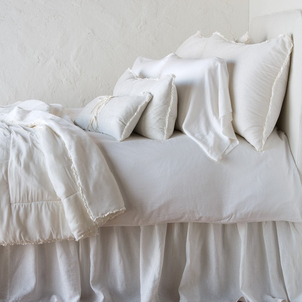 Carmen Throw Pillow | Winter White | Silk velvet lumbar pillow with petite ruffle on a monochromatic, neatly made bed with matching accessory pieces - side view.