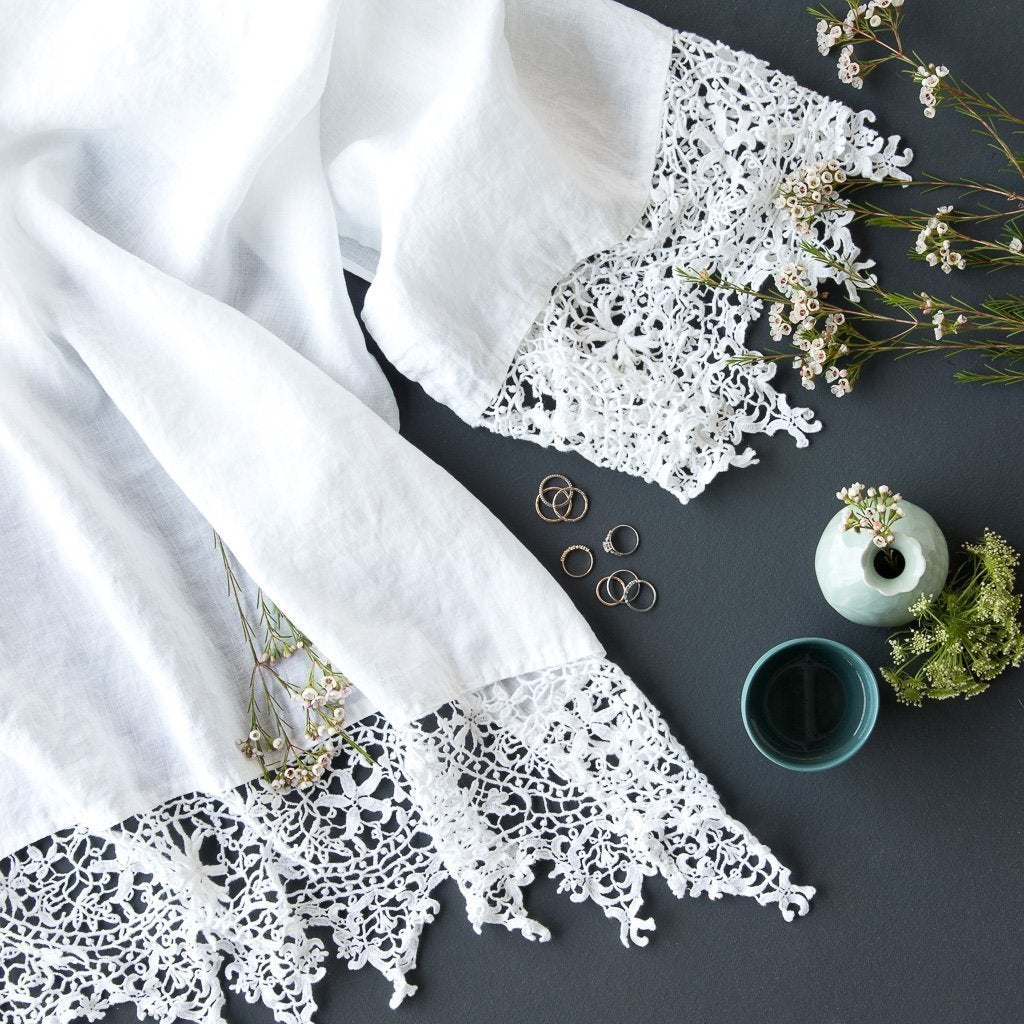 White | Frida guest towel in white draped on a dark background with blue green vases and delicate flowers - overhead view.