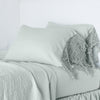 Frida Pillowcase (Single) | Eucalyptus | Lace trimmed linen pillowcases leaning against a plain wall on a monochromatic bed - cropped three-quarter angle.