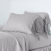 Frida Pillowcase (Single) | Fog | Lace trimmed linen pillowcases leaning against a plain wall on a monochromatic bed - cropped three-quarter angle.