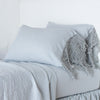 Frida Pillowcase (Single) | Mineral | Lace trimmed linen pillowcases leaning against a plain wall on a monochromatic bed - cropped three-quarter angle.