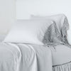 Frida Pillowcase (Single) | Sterling | Lace trimmed linen pillowcases leaning against a plain wall on a monochromatic bed - cropped three-quarter angle.