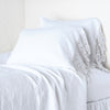 Frida Pillowcase (Single) | White | Lace trimmed linen pillowcases leaning against a plain wall on a monochromatic white bed - cropped three-quarter angle.
