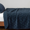 Harlow Coverlet | Midnight | Quilted cotton velvet coverlet, with top folded back to showcase linen back, draped over a white fitted sheet - side view.